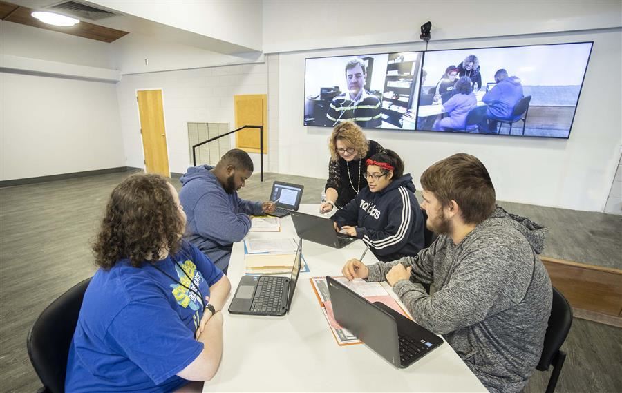 A teacher stands with 4 students sit around a table with their laptops. An RBT Zooms on a screen. 